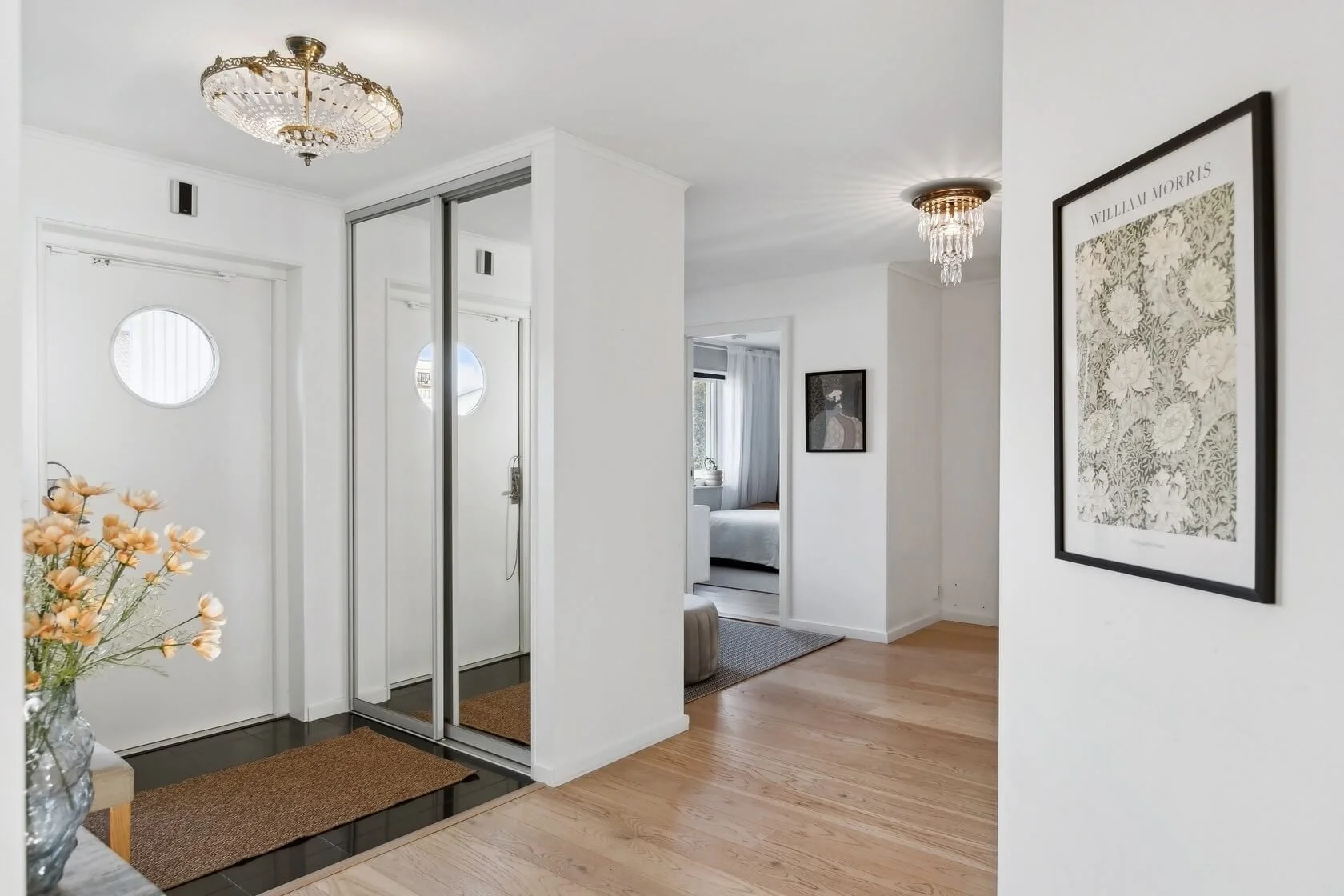 Bright apartment entryway with white walls, hardwood floors, a chandelier, a flower vase, a mirror on a closet door, and framed artwork.