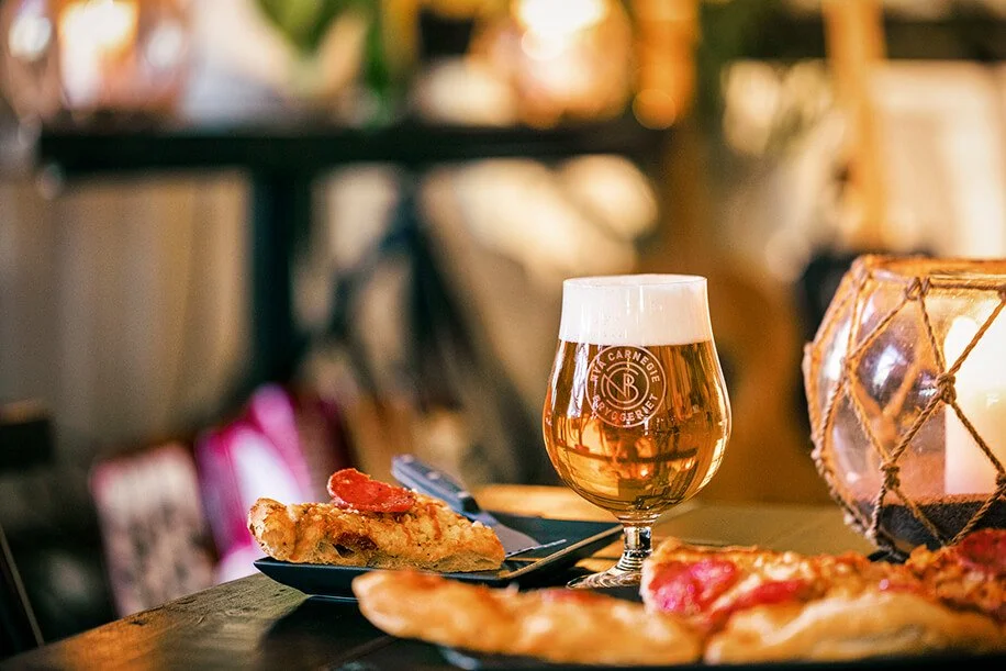 A glass of beer with a foamy head and slice of pepperoni pizza on a plate on a table in a cozy setting.