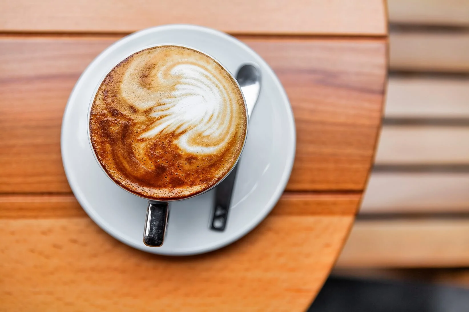 A cup of coffee with latte art on top, served on a white saucer with a spoon, placed on a wooden table.