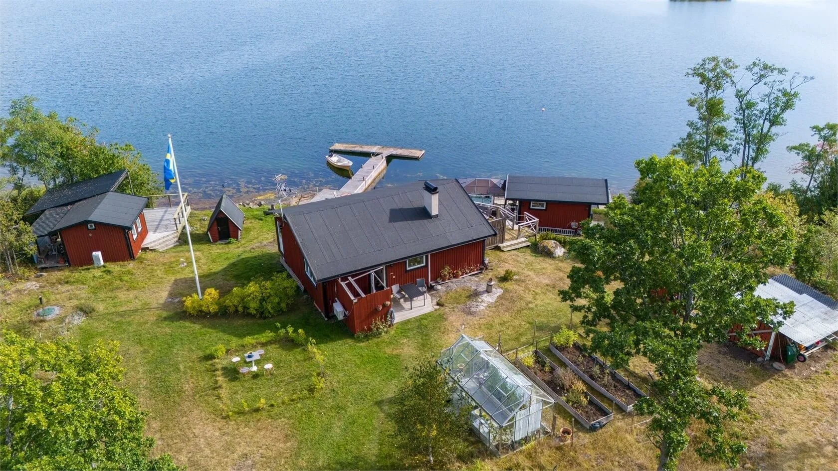 Aerial view of red wooden houses next to a lake with a dock and boat, surrounded by green trees and lawn.