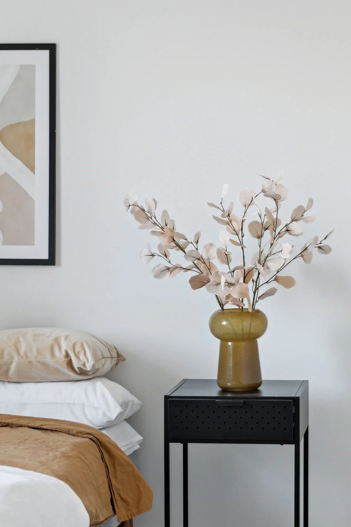 A bedroom with a black side table holding a yellow vase with white and beige artificial flowers, a portion of a bed with beige and white pillows, and a framed abstract art piece on a light gray wall.