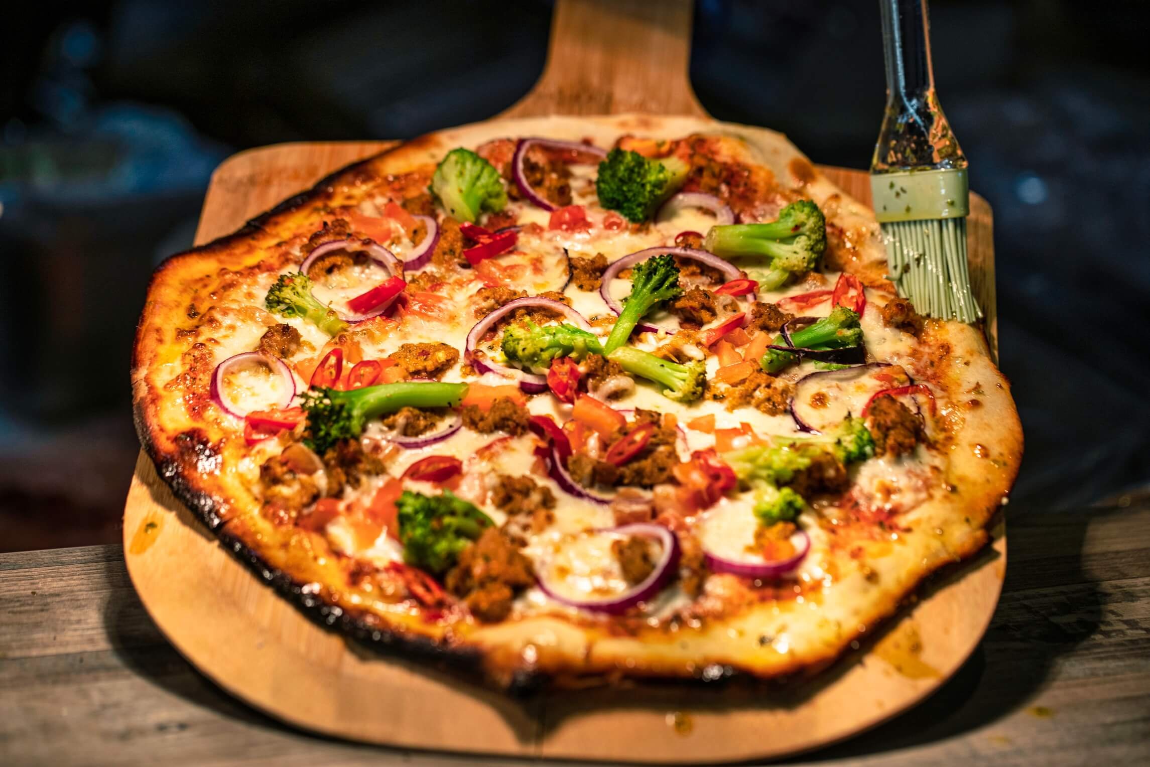 A pizza with broccoli, red onions, ground beef, and red chili peppers on a wooden pizza peel.