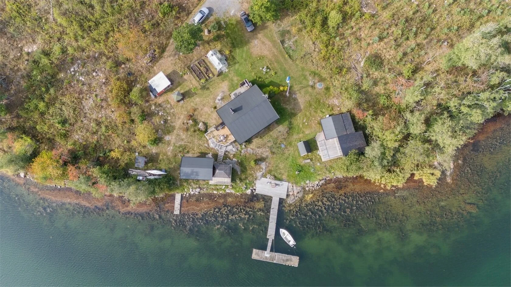 Aerial view of a lakeside property with multiple buildings, a dock with a boat, surrounded by trees, and a grassy yard.