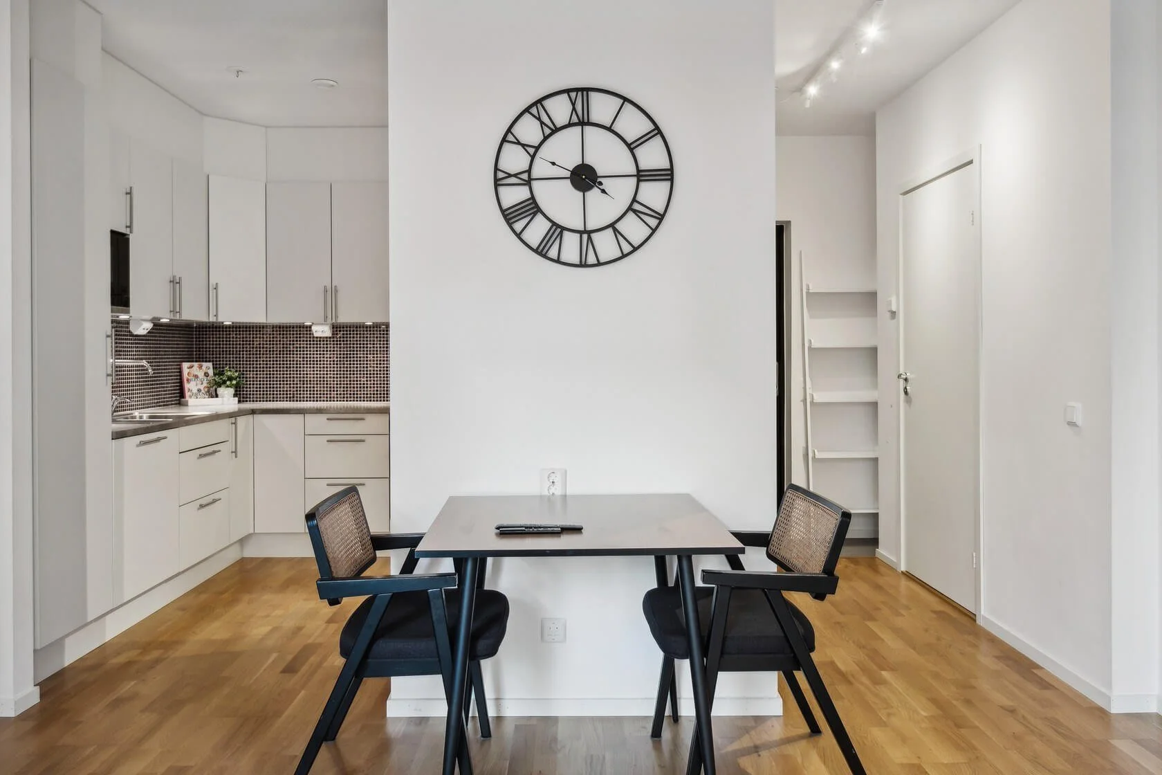 Modern dining area with a black clock on white wall, a table with two chairs, and a kitchen in the background.
