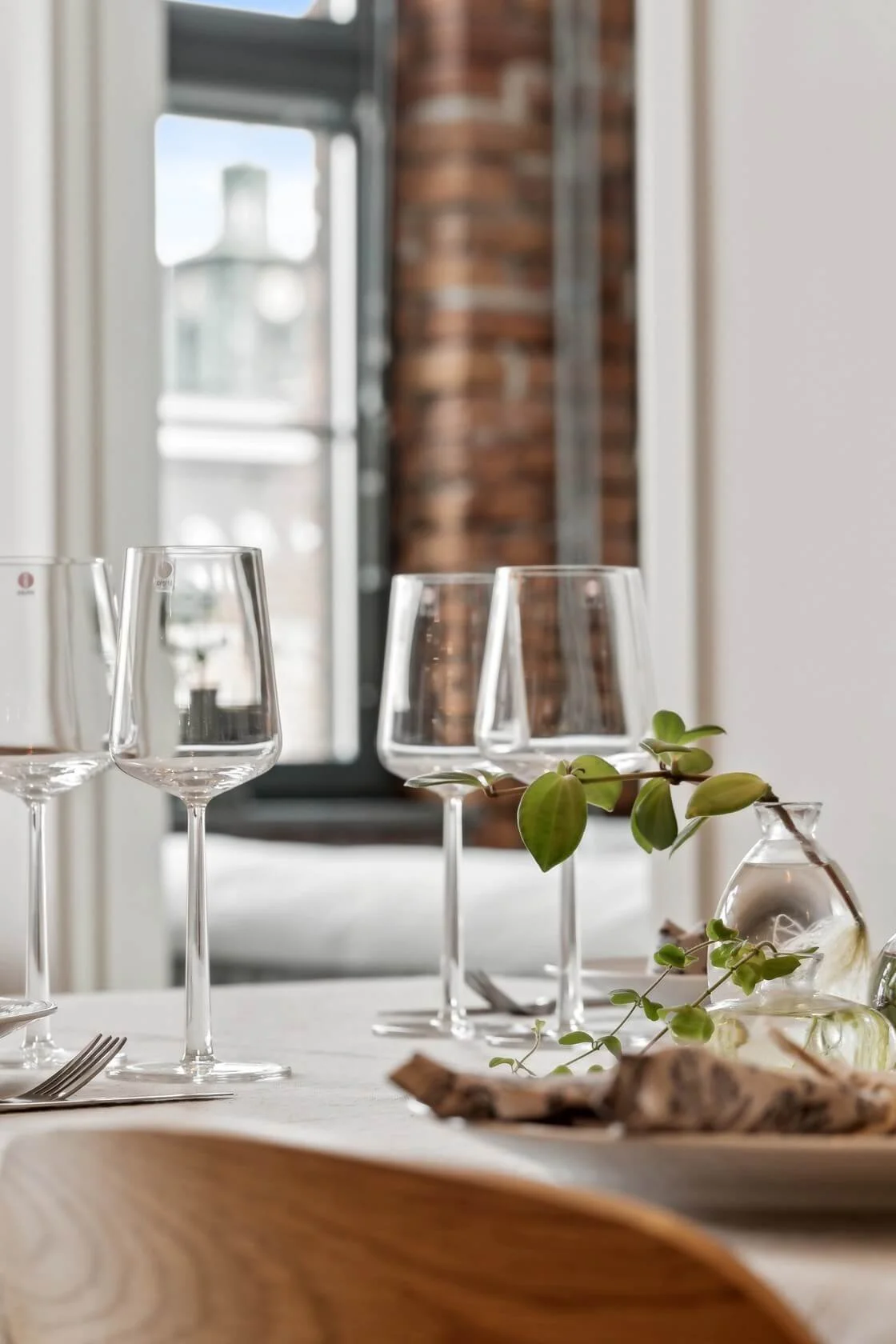 A table set for a meal with four empty wine glasses, a small leafy green plant in a glass vase, and some folded napkins on a light-colored tablecloth, with a background of a window and brick wall.