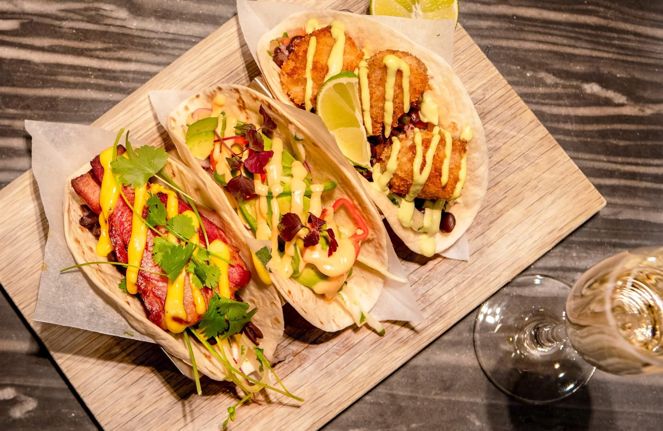 Three tacos topped with sauces, cilantro, and vegetables on a wooden serving board, with a glass of white wine nearby.