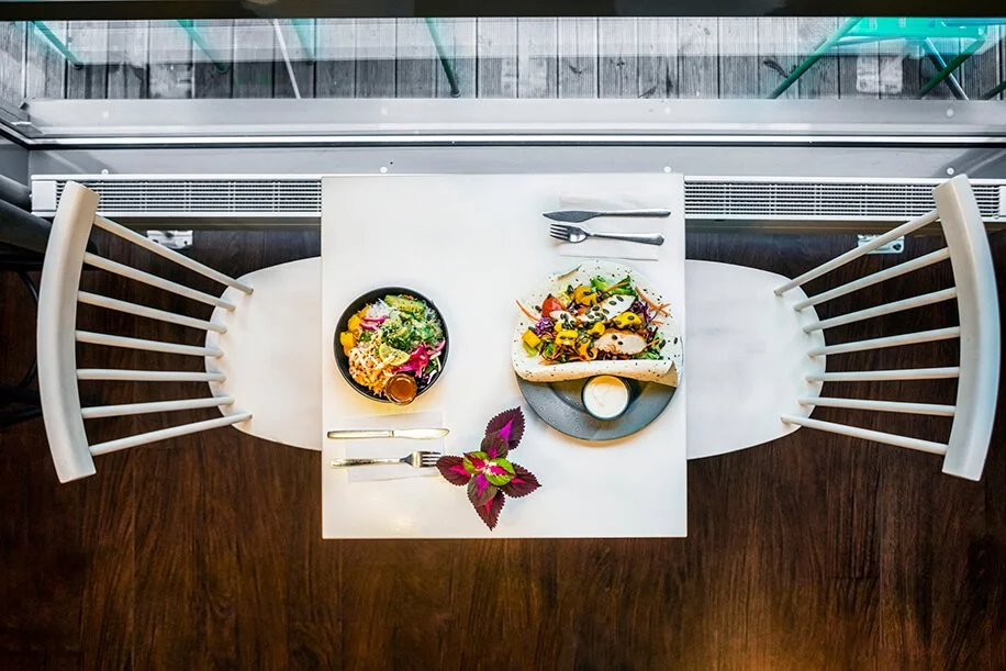 A table set for a meal with two plates of food, salad, and utensils, viewed from above near a window with a balcony.