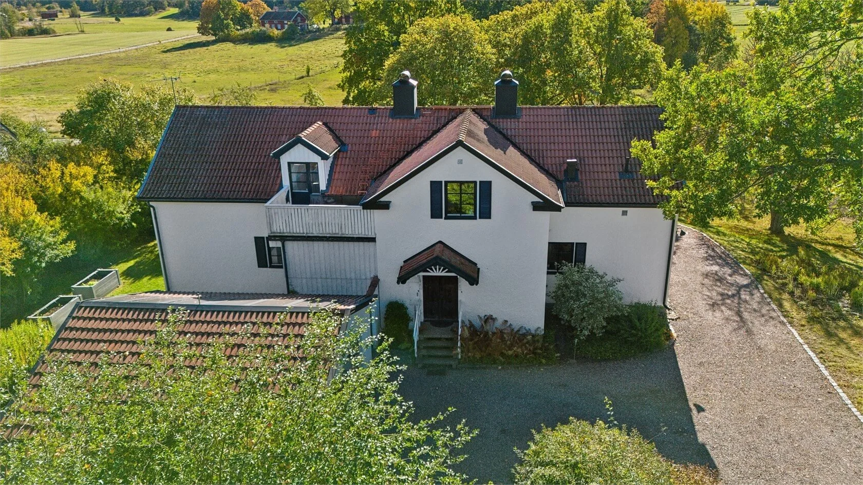 Aerial view of a white house with a red-tiled roof and black shutters, surrounded by trees and green landscape.