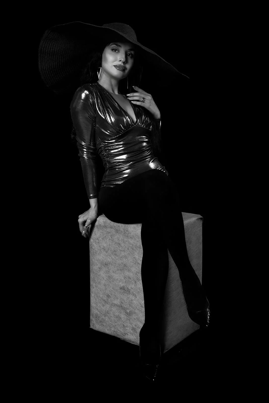 A woman in a shiny, tight black dress and high heels, wearing a large sunhat, sitting on a cube in a black and white photograph.