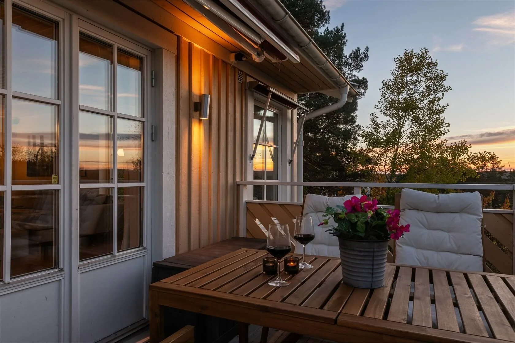 Balcony at sunset with a wooden table, two glasses of red wine, a candle, a flowerpot with pink flowers, and cushioned chairs.