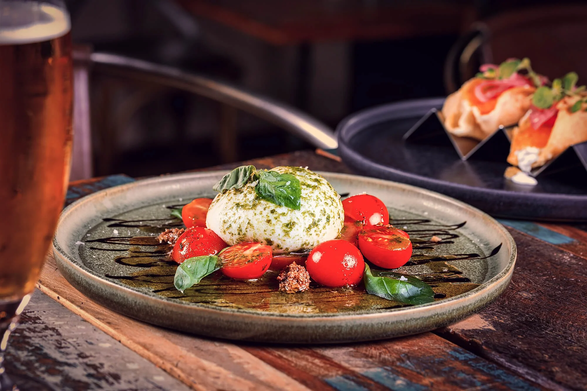 A plate with a serving of burrata cheese topped with green pesto, surrounded by cherry tomatoes, basil leaves, and balsamic glaze drizzle, on a rustic wooden table. In the background, a black plate with assorted mini sandwiches or tacos, and a copper