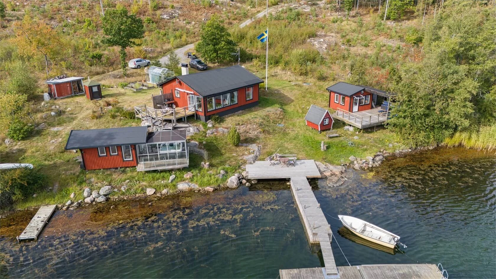 Aerial view of a lakeside property with three red cabins, a small red shed, a boat dock with a rowboat, and a grassy yard with trees and a Swedish flag on a flagpole.