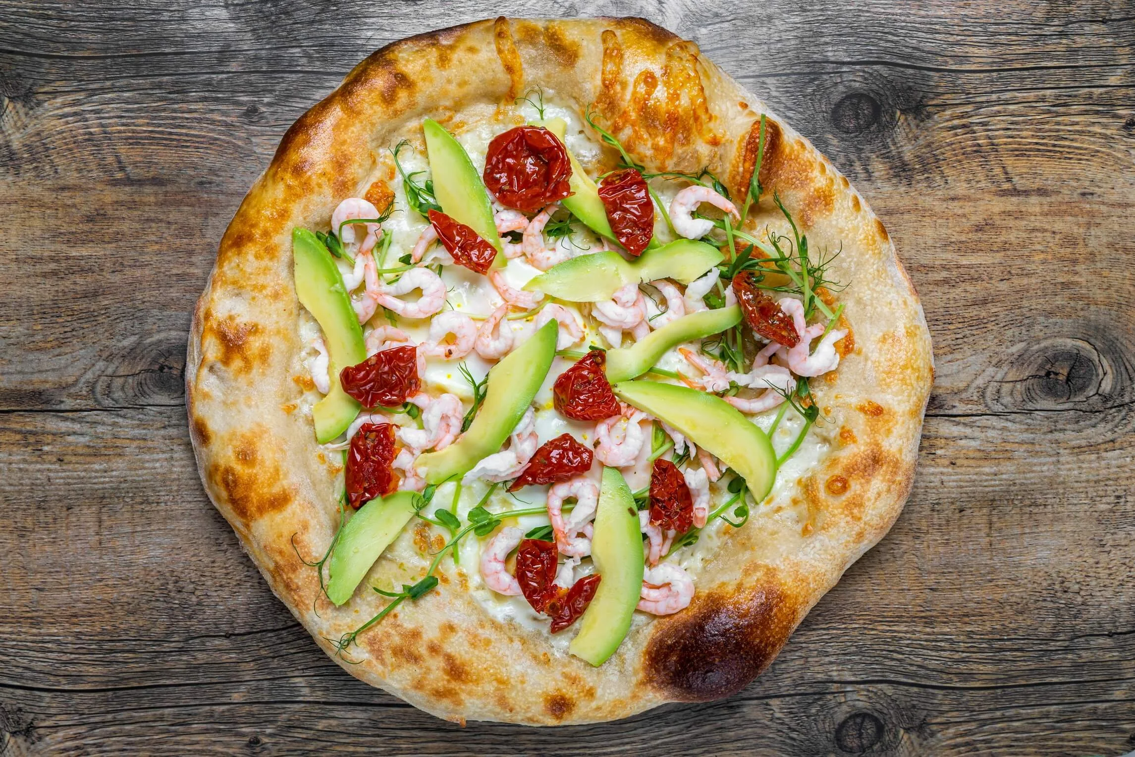 A seafood pizza with shrimp, sun-dried tomatoes, avocado slices, and greens on a wooden surface.