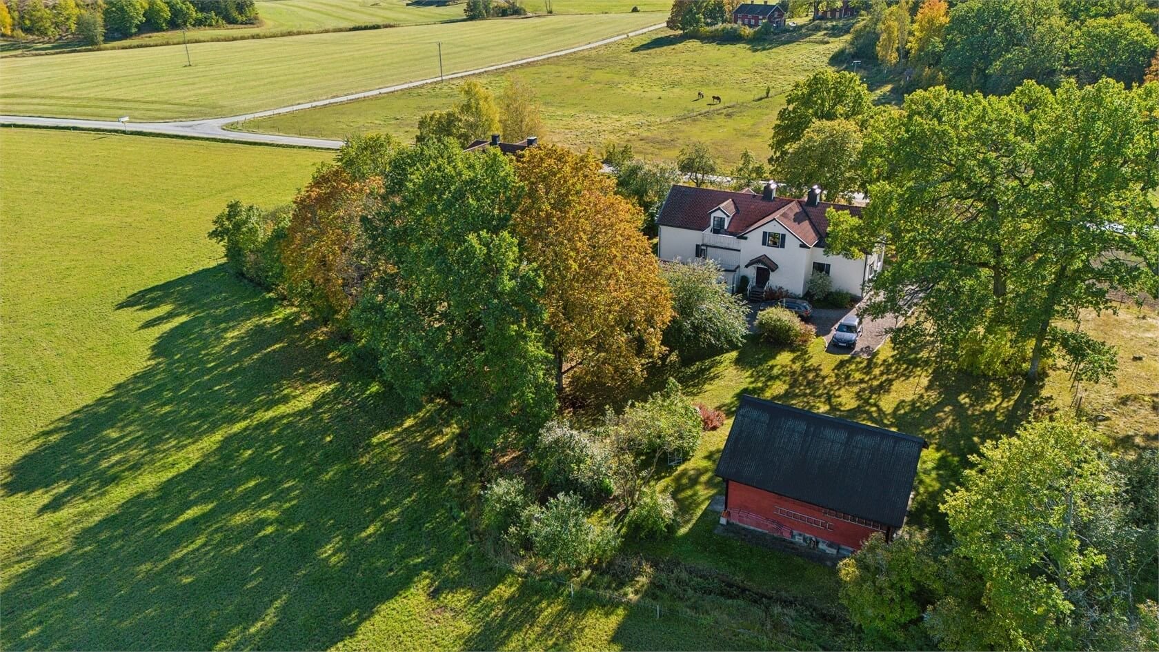 Aerial view of a white house with a red roof, surrounded by large green trees and a well-maintained lawn. A gravel driveway with parked cars leads to the house, and there is a red barn nearby. Horses can be seen grazing in the background on open past