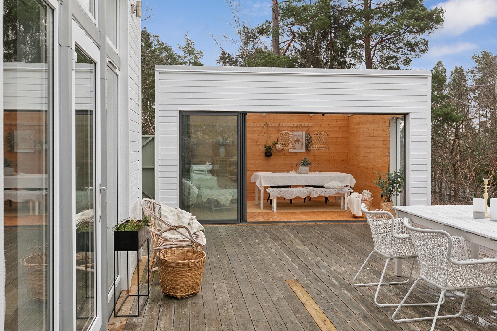 A deck with outdoor furniture, including a white table and wicker chairs, leading to a cozy wooden indoor space with a table, benches, and wall decorations, surrounded by trees under a partly cloudy sky.