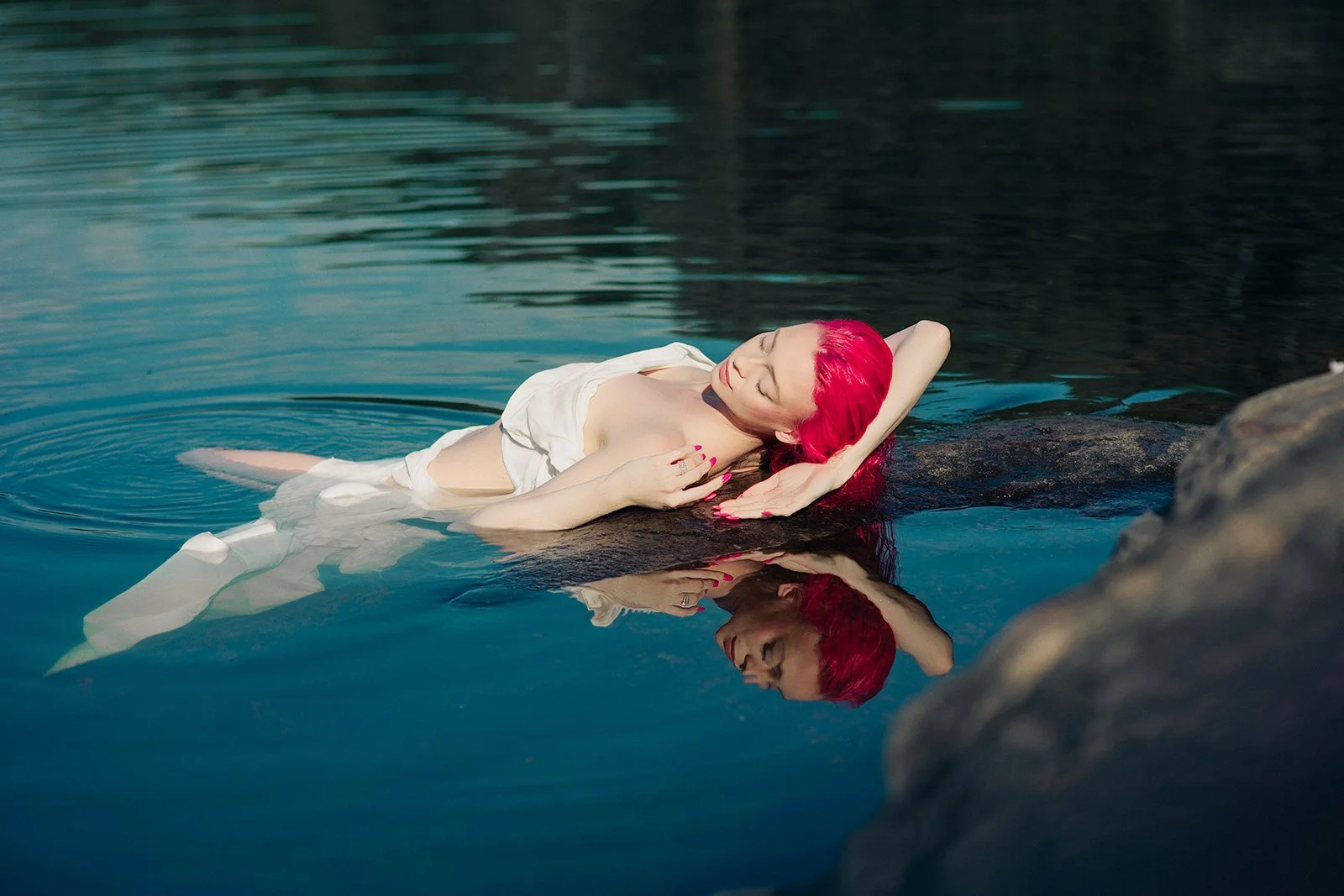 Woman with bright red hair reclining on a rock in a body of water with calm ripples, wearing a white dress with her eyes closed and one arm resting behind her head.