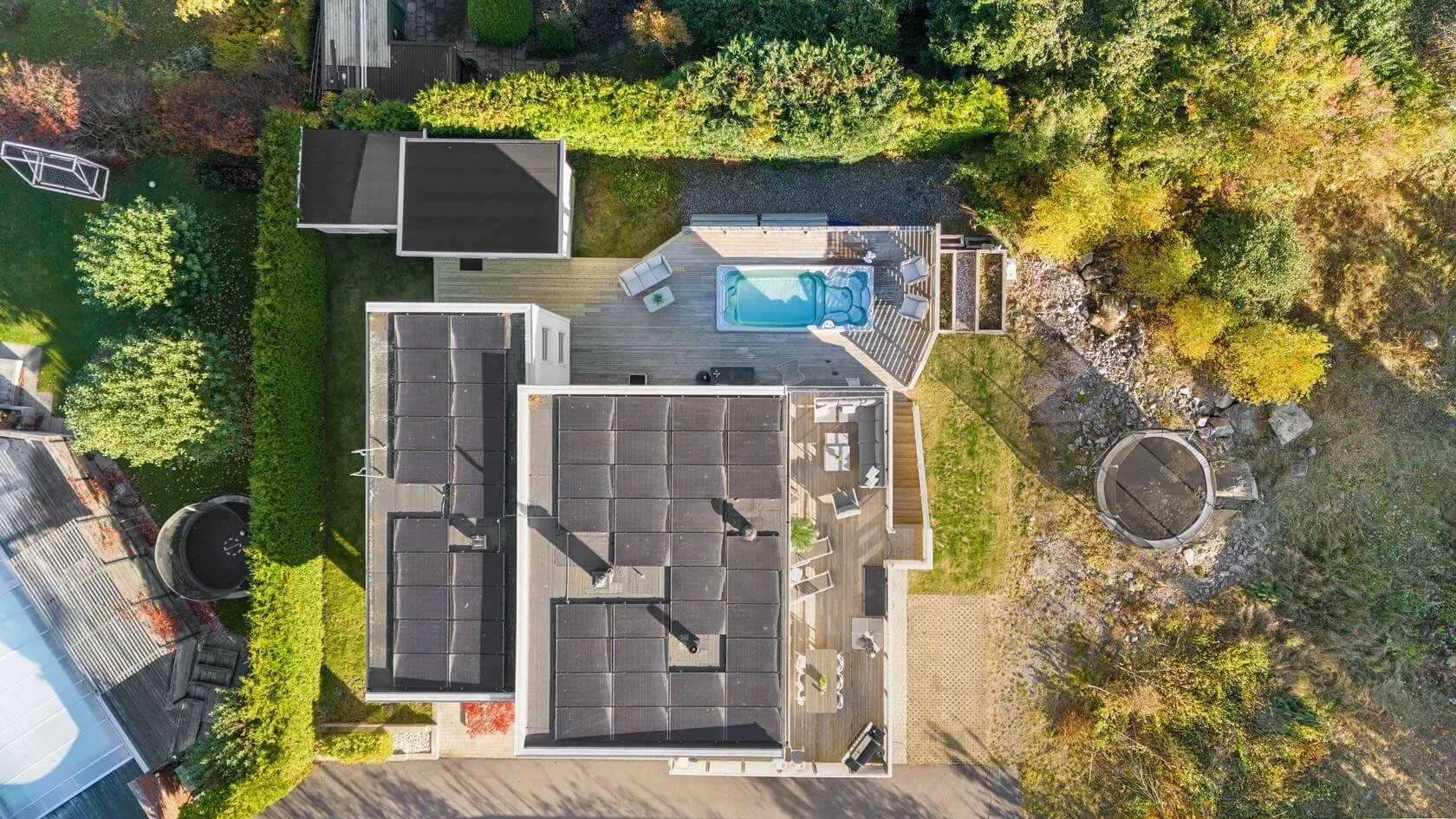Aerial view of a backyard with a house, a deck with outdoor furniture, a swimming pool with a slide, a trampoline, and surrounding lush greenery and trees.
