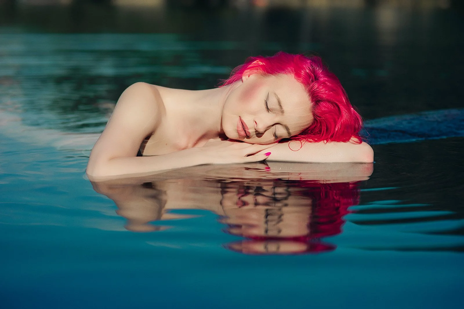 A woman with bright red hair laying on her side in water with her eyes closed, resting her head on her arm.
