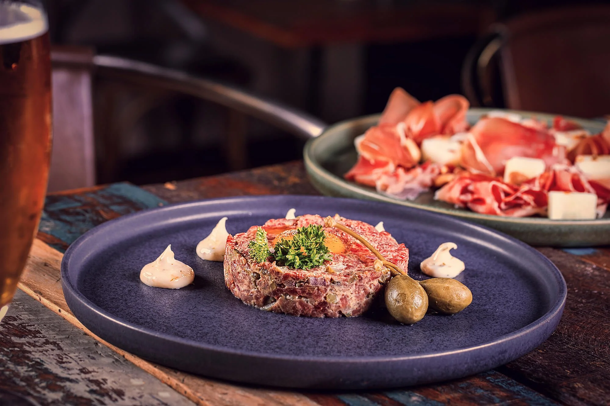 Raw beef steak tartare garnished with herbs, served with dollops of mayonnaise and caperberries on a dark blue plate, with a plate of sliced cured meats and cheese in the background.