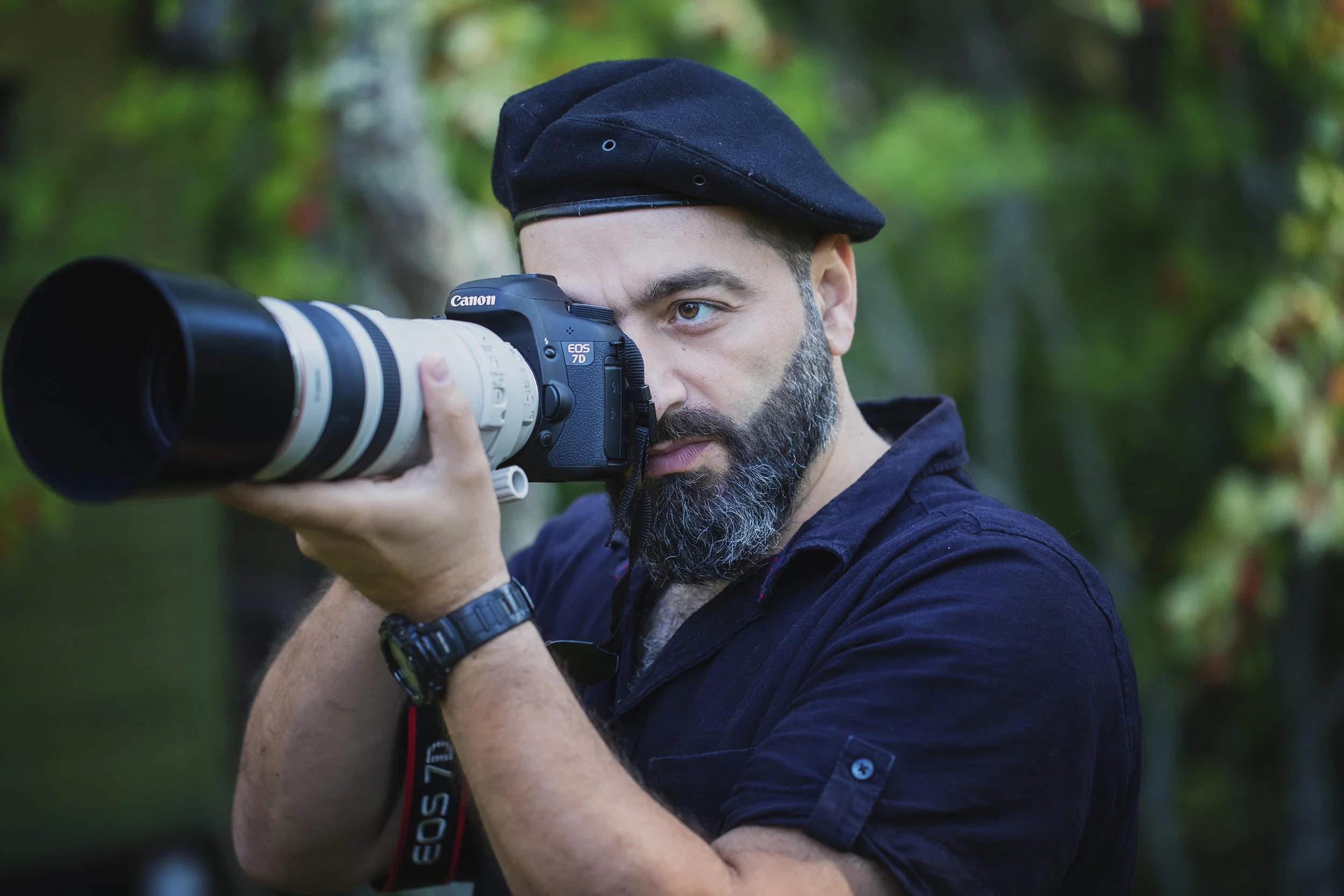 Man with a full beard wearing a black beret and black shirt, holding a Canon EOS 7D camera with a large telephoto lens, aiming it through a forest setting.