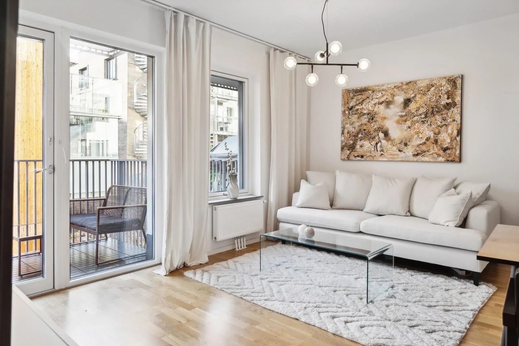 Living room with white sofa, glass coffee table, beige painting on wall, wooden floor, white curtains, and balcony with outdoor chair.