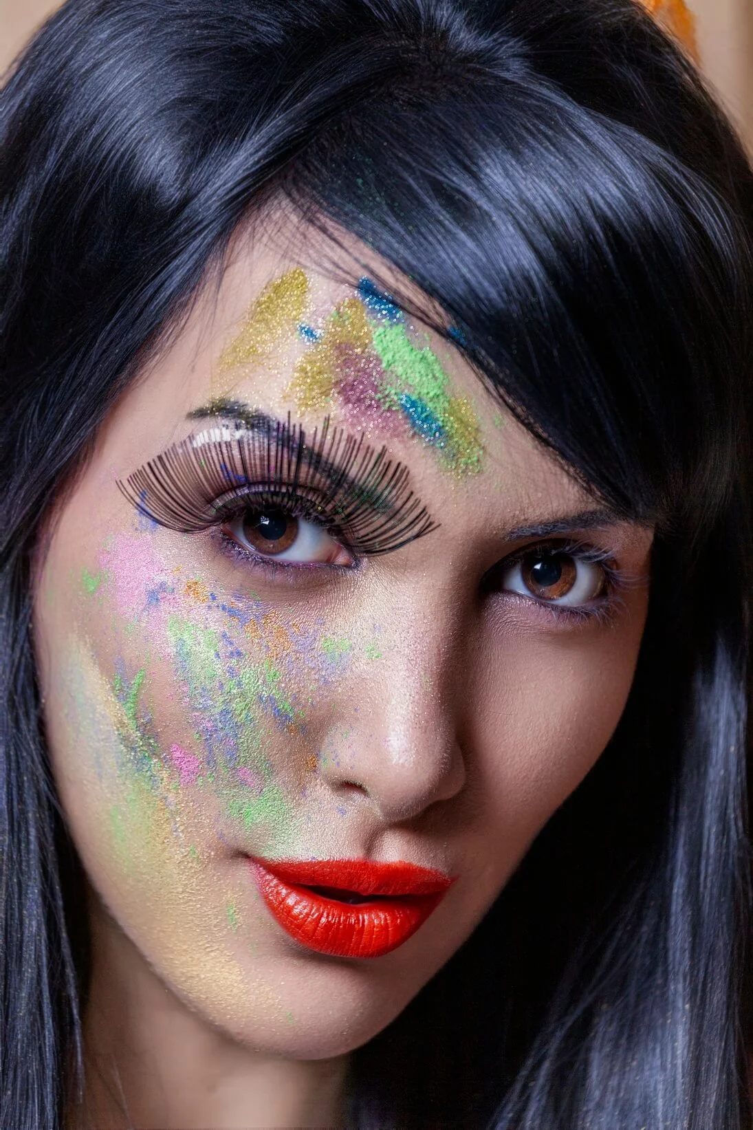 A woman with long dark hair, colorful glitter makeup on her face, large false eyelashes, and bright red lipstick.