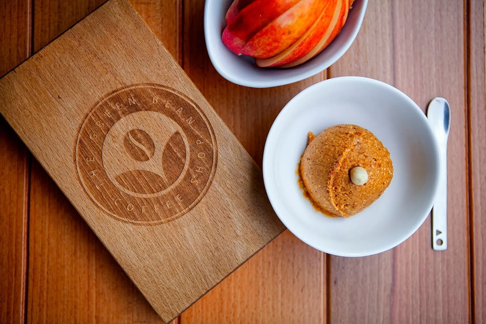 A white bowl with an ice cream scoop topped with a white chocolate chip, a small white bowl with sliced peaches, a wooden box with a logo, and a spoon, all on a wooden table.