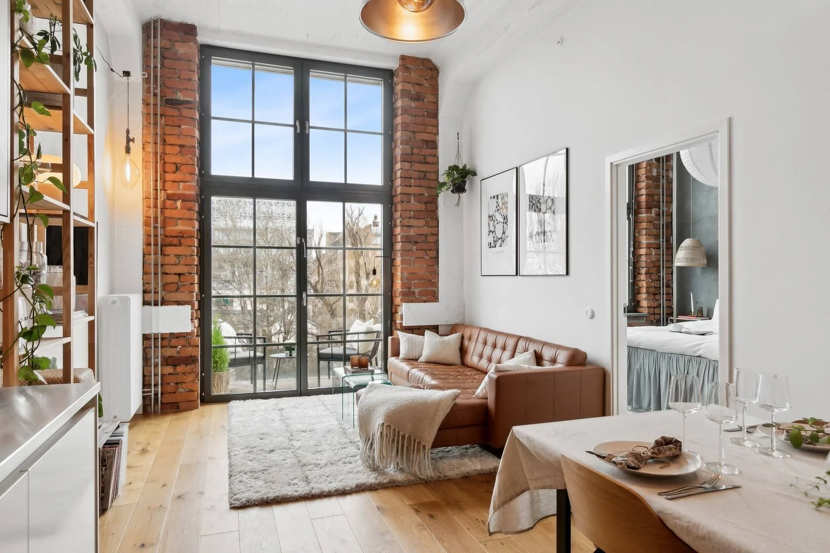 Living room with large window, white walls, wood flooring, a brown tufted leather sofa with pillows and blanket, glass coffee table, wall art, and balcony doors leading outside.