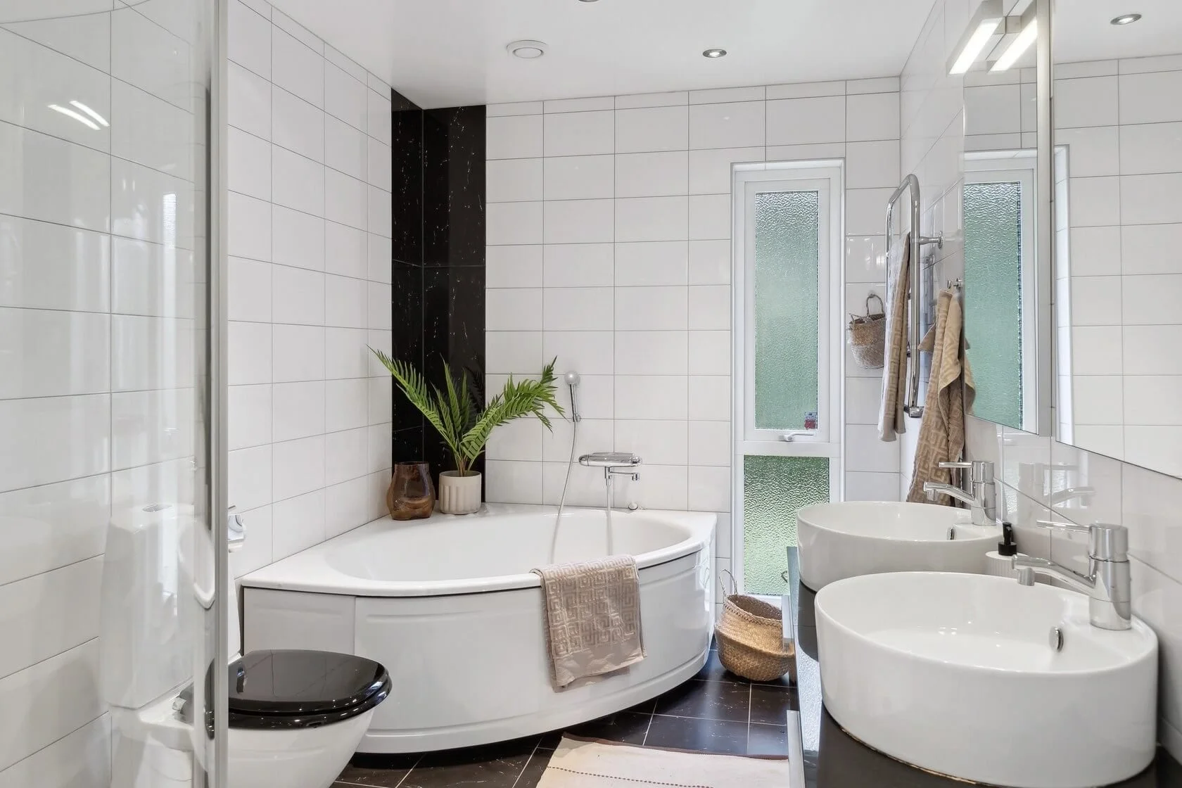 Modern bathroom with white tiled walls, black floor, oval bathtub, two white sinks, a toilet, a window, a mirror, and decor including plants and baskets.