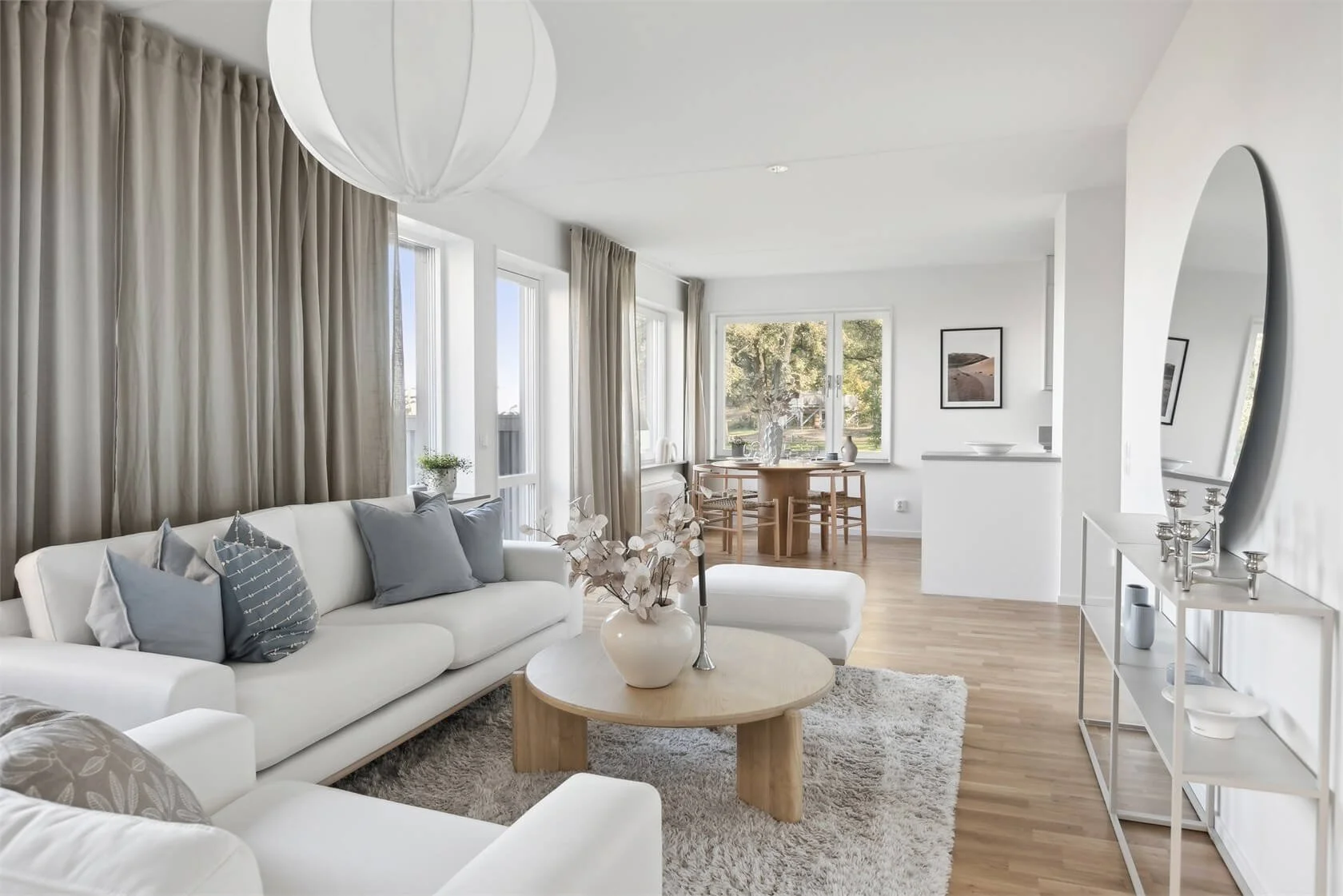 Bright living room with white sofas, beige curtains, round coffee table with white vase and dried flowers, and a dining area with wooden table and chairs in the background, large windows showing greenery.