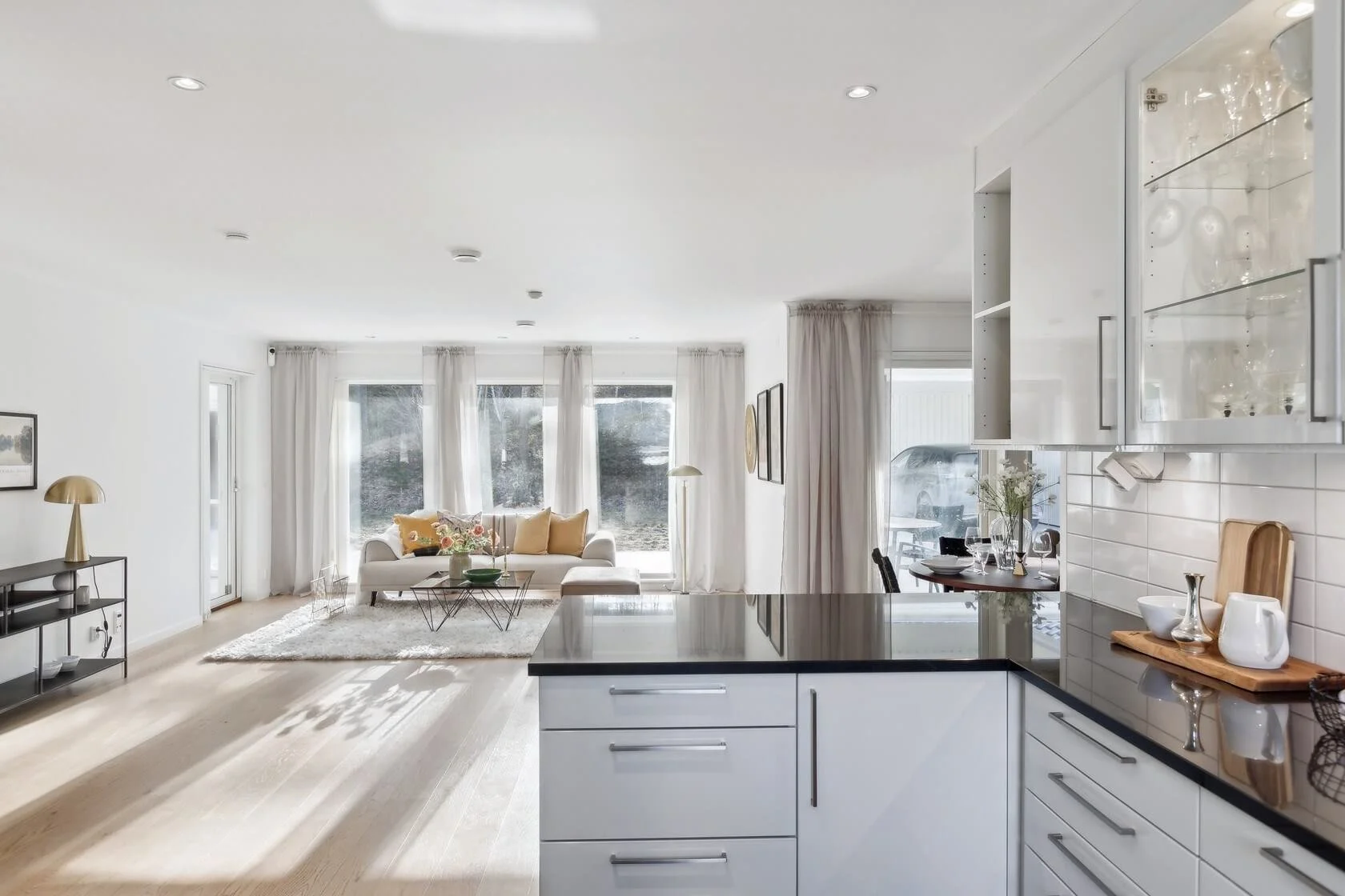 Modern open-concept living room and kitchen with white walls, hardwood flooring, large windows with white curtains, a white sofa with beige pillows, a coffee table with decorative items, a black console with a gold lamp, and a kitchen counter with wh