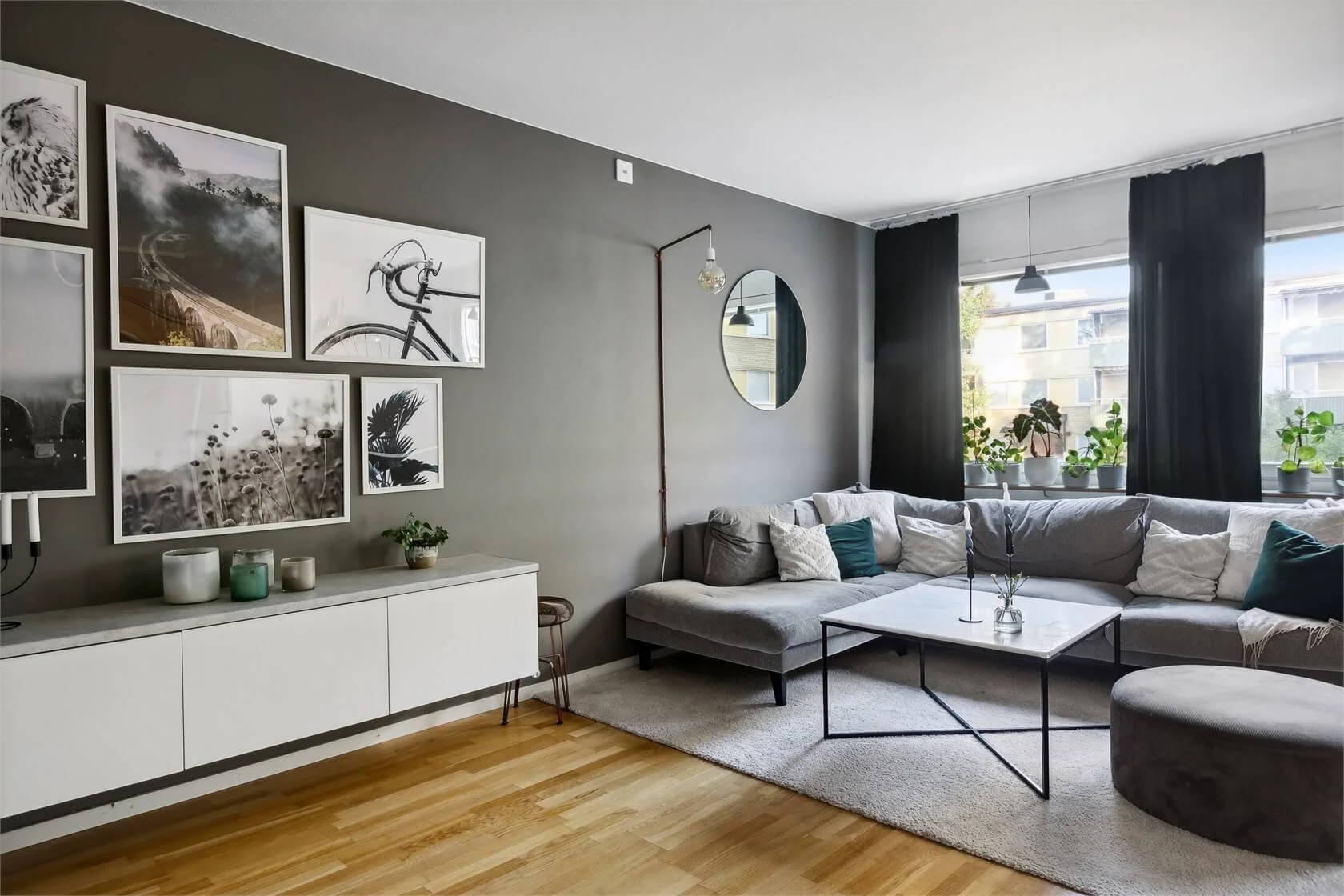 Modern living room with a gray wall, black and white artwork, a gray sectional sofa with pillows, a white coffee table, and a row of potted plants on the window sill.