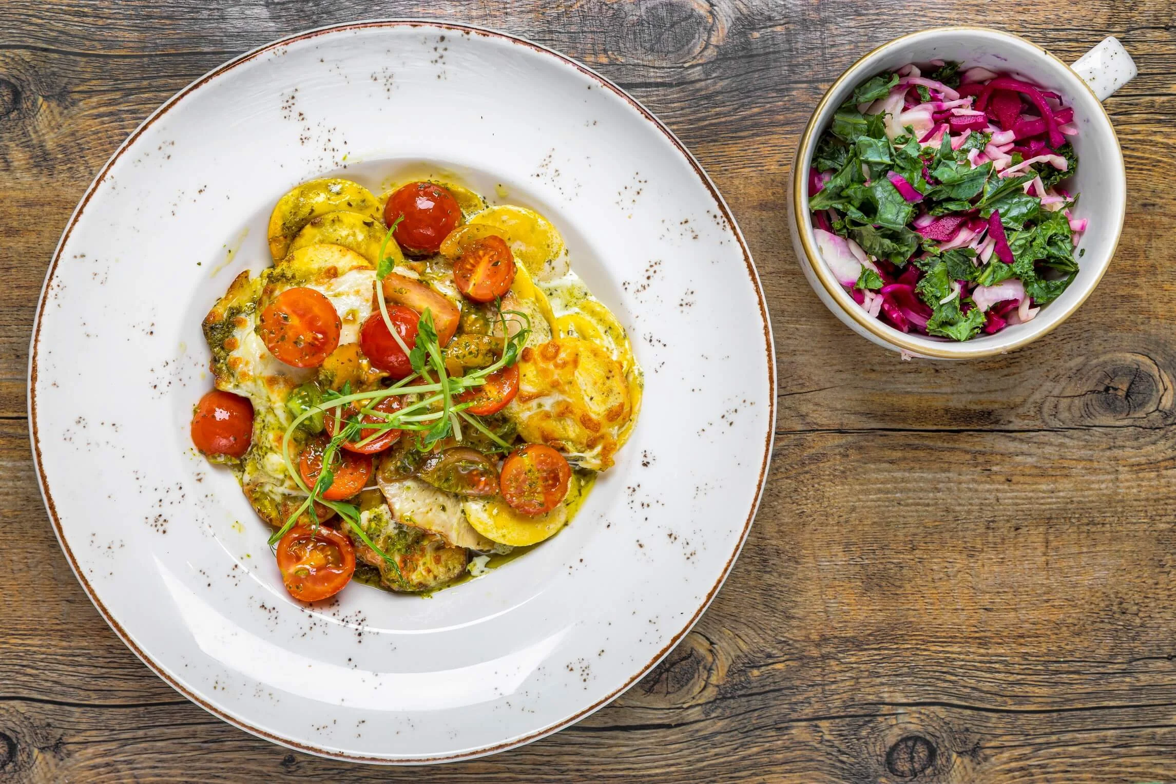 Plate of roasted vegetables with cherry tomatoes and microgreens, served on a rustic wooden table, alongside a bowl of colorful shredded cabbage and greens salad.