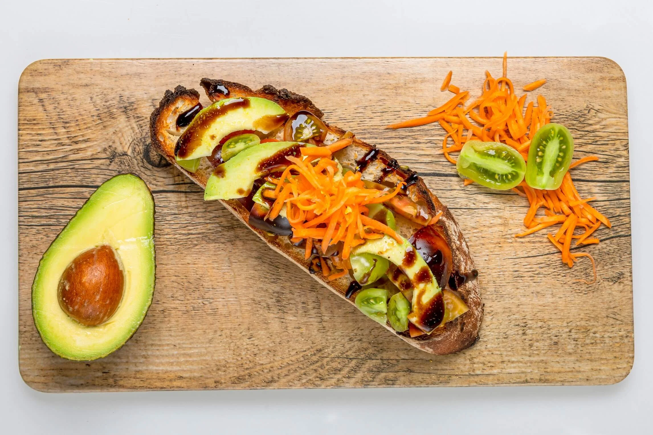 Open-faced sandwich on toasted bread with avocado slices, shredded carrots, green and purple olives, drizzled with balsamic glaze, served with half an avocado and shredded carrots with green olives on a wooden cutting board.