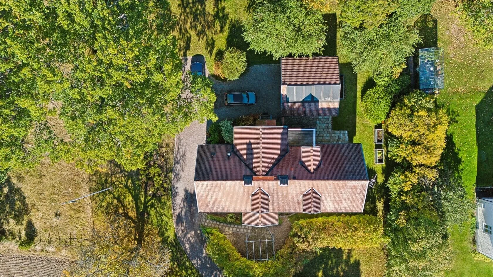 Aerial view of a house with a red tiled roof, surrounded by green trees, a driveway with cars, and a landscaped yard with garden beds.