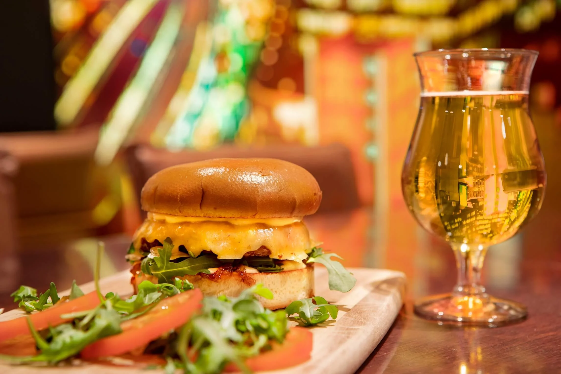 A cheeseburger with a bun, cheese, lettuce, and tomato slices next to a glass of beer, on a wooden table with colorful blurry background.