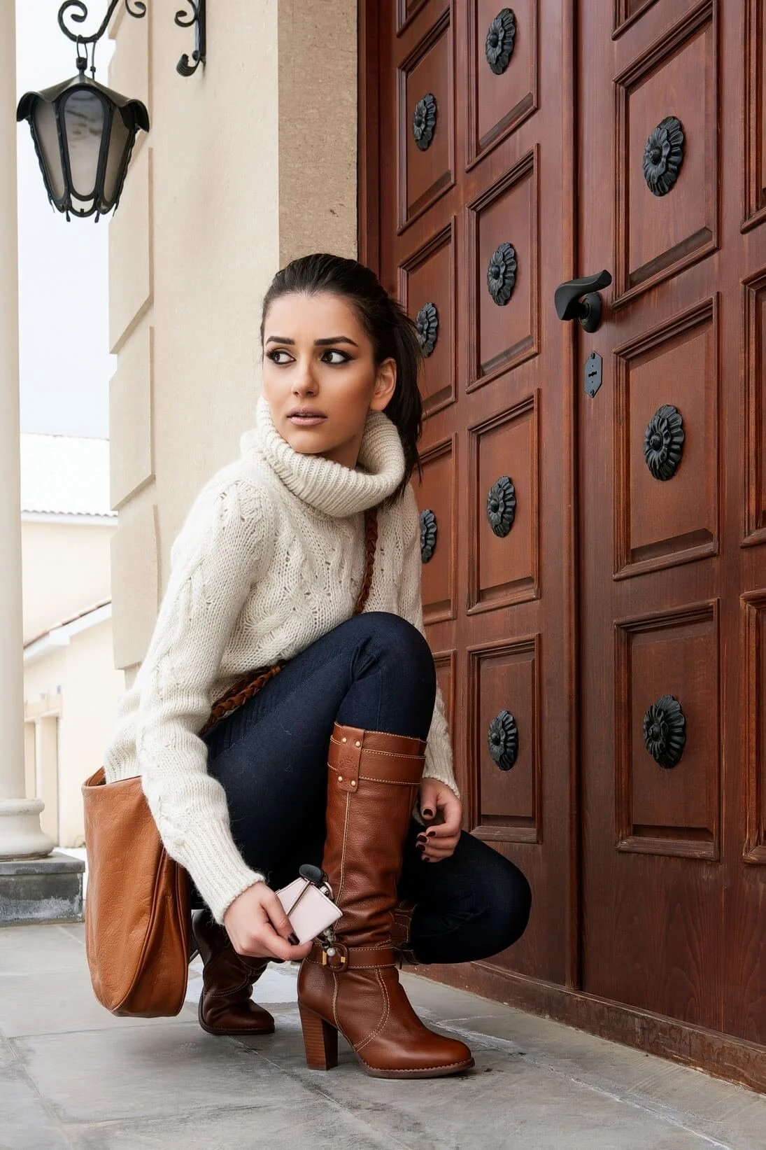 A woman crouching on the porch in front of a wooden door, wearing a cream sweater, dark jeans, and brown knee-high boots, holding a small device and carrying a brown shoulder bag.