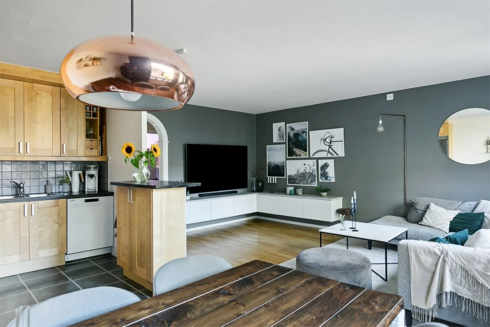 Modern living room with gray walls, a flat-screen TV, framed black and white photos, a white TV stand, sectional sofa with cushions, a coffee table, and a large mirror on the wall. Kitchen area with wooden cabinets, black countertop, sunflower vase, 