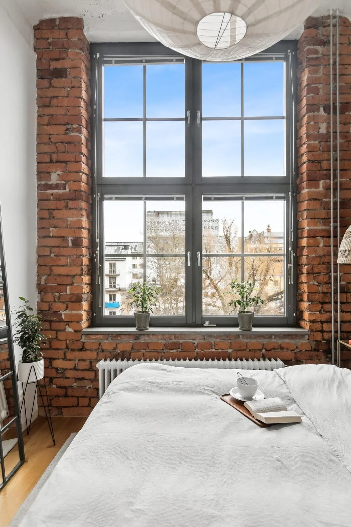 A bedroom with a large window framed by red brick walls, two potted plants on the windowsill, a bed with white bedding, a tray with a cup and an open book, and a paper lantern ceiling light.