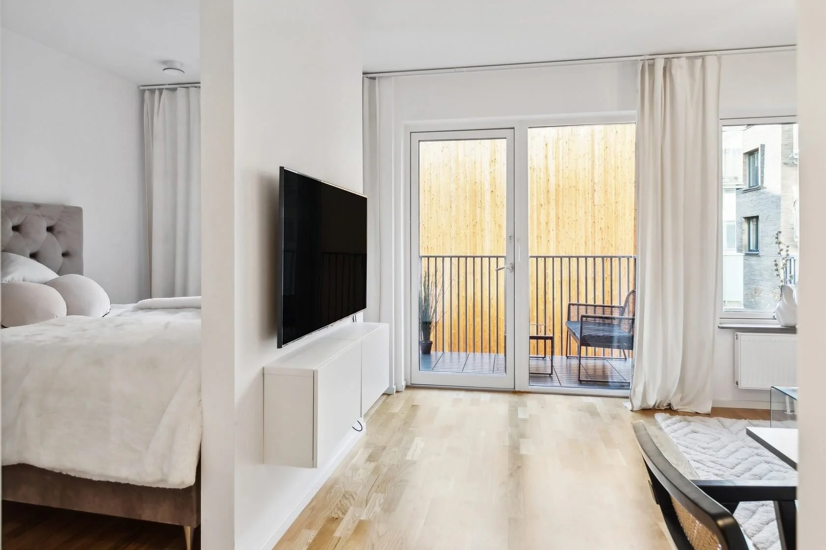 Living room with a bed partially visible on the left, a wall-mounted TV, and a balcony with a wooden fence and outdoor furniture.