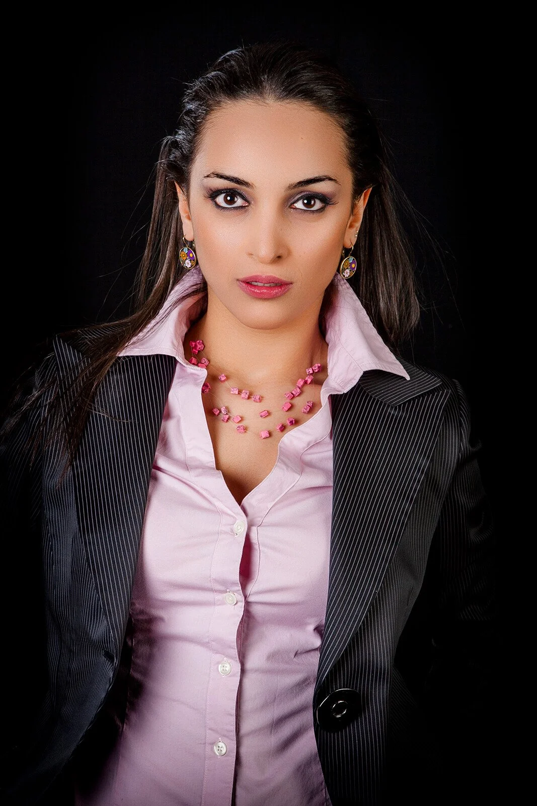 A woman in business attire with dark hair, wearing earrings and a pink necklace against a black background.
