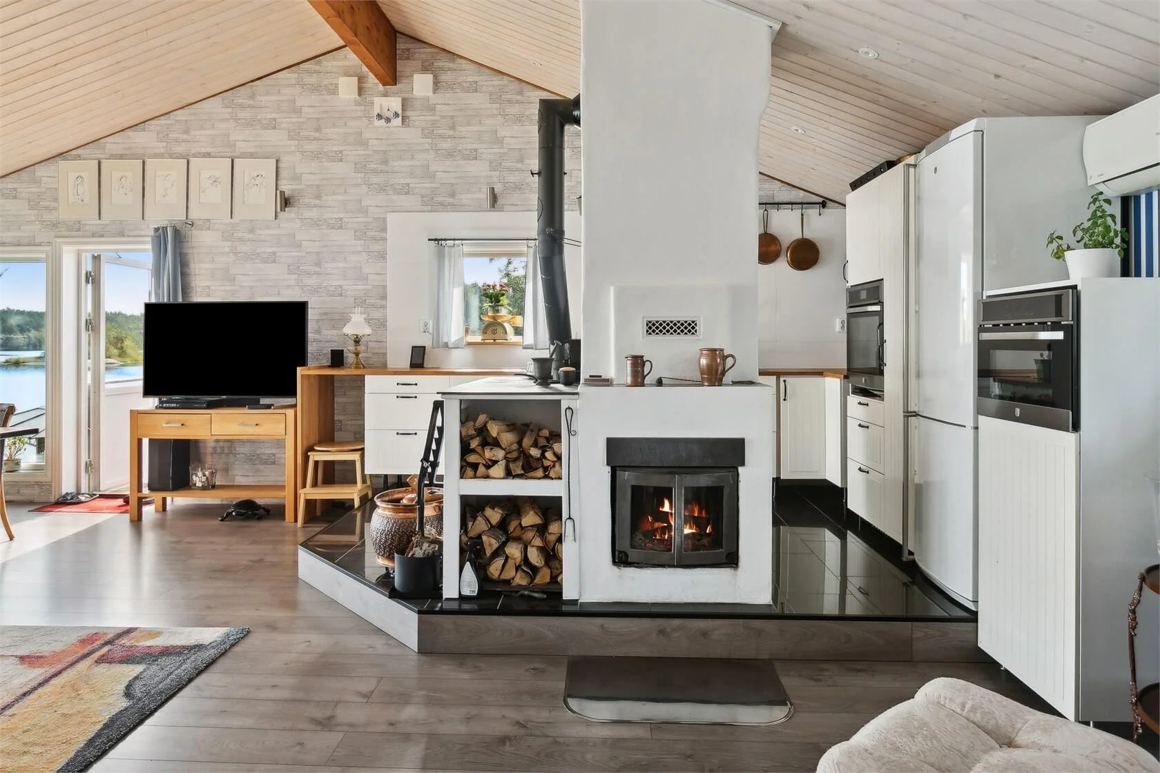 Cozy living room and kitchen with wood vaulted ceiling, fireplace, television, and lake view through glass doors.