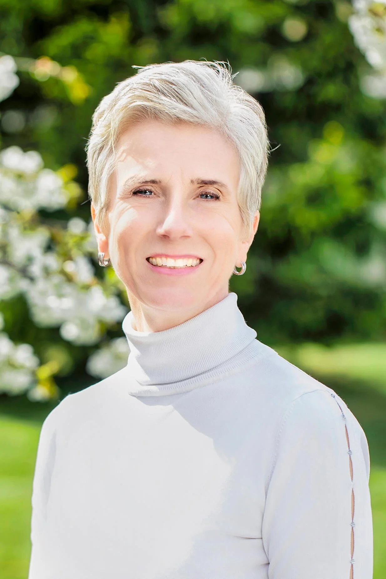 A smiling woman with short gray hair, wearing a white turtleneck sweater, standing outdoors with greenery in the background.