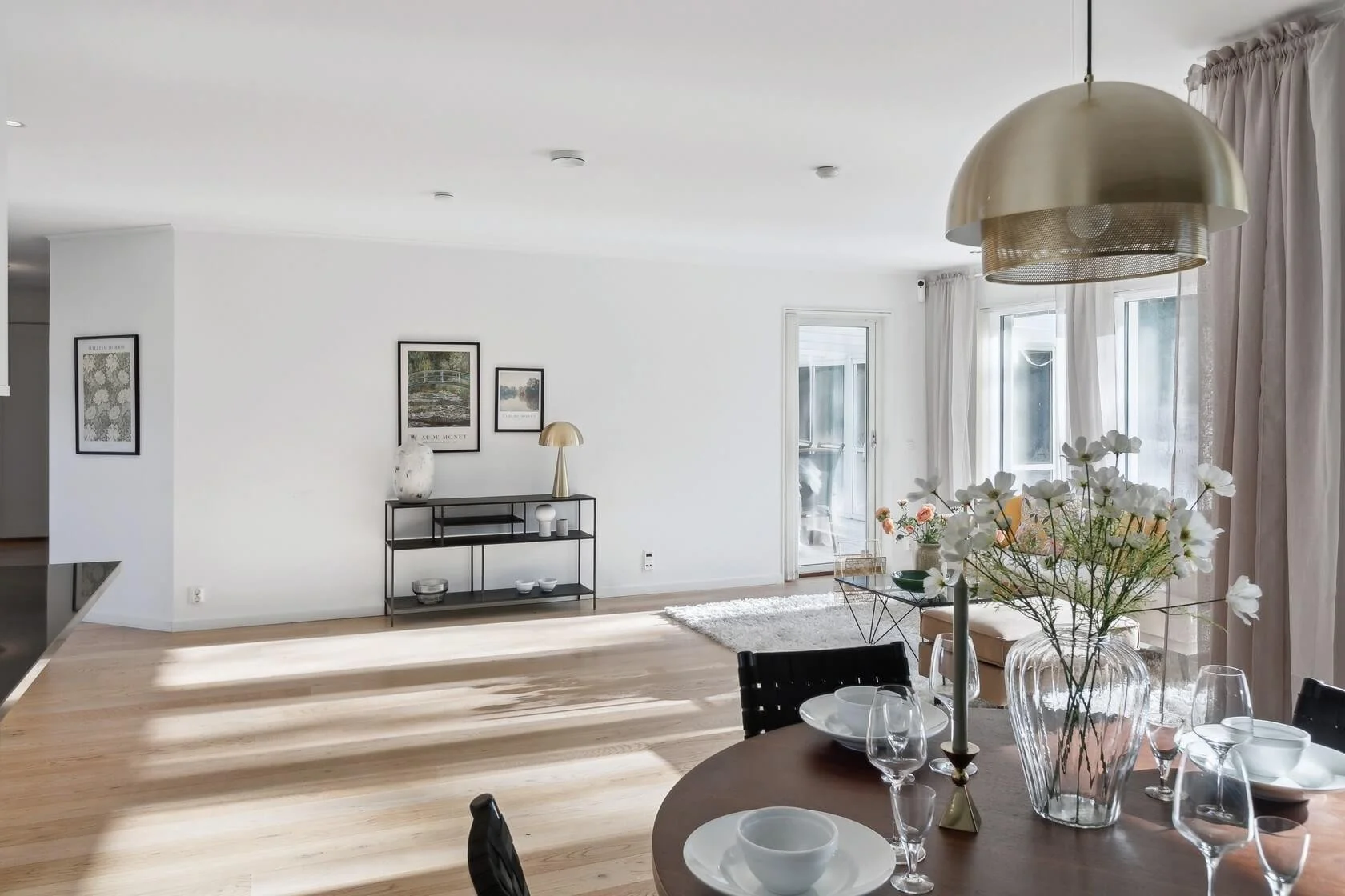 An open living and dining area with a wooden floor, white walls, and large windows with sheer curtains. The dining table has a centerpiece of white flowers in a glass vase, surrounded by plates, wine glasses, and silverware. A chandelier hangs above 