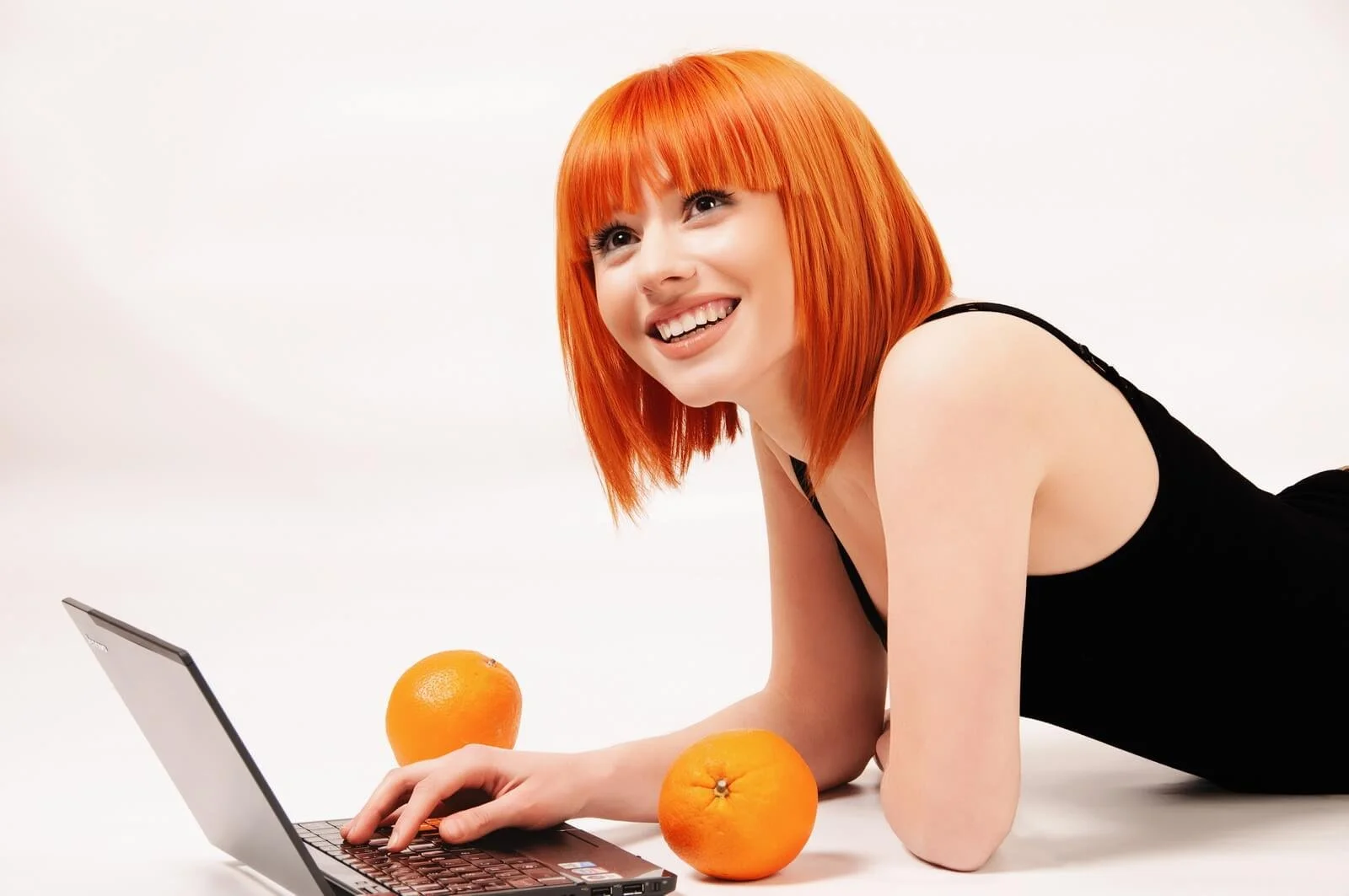 A woman with red hair lying on the floor, smiling, using a laptop, with two oranges placed nearby.