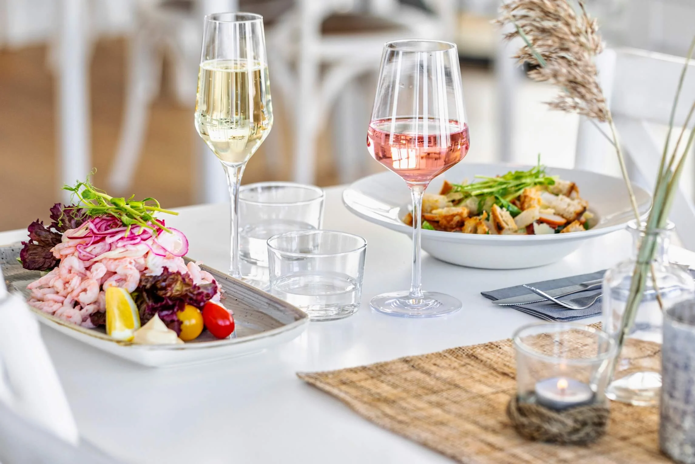 A table set for dining with two glasses of wine, one white and one rosé, along with a salad, appetizers, and decorative flowers.