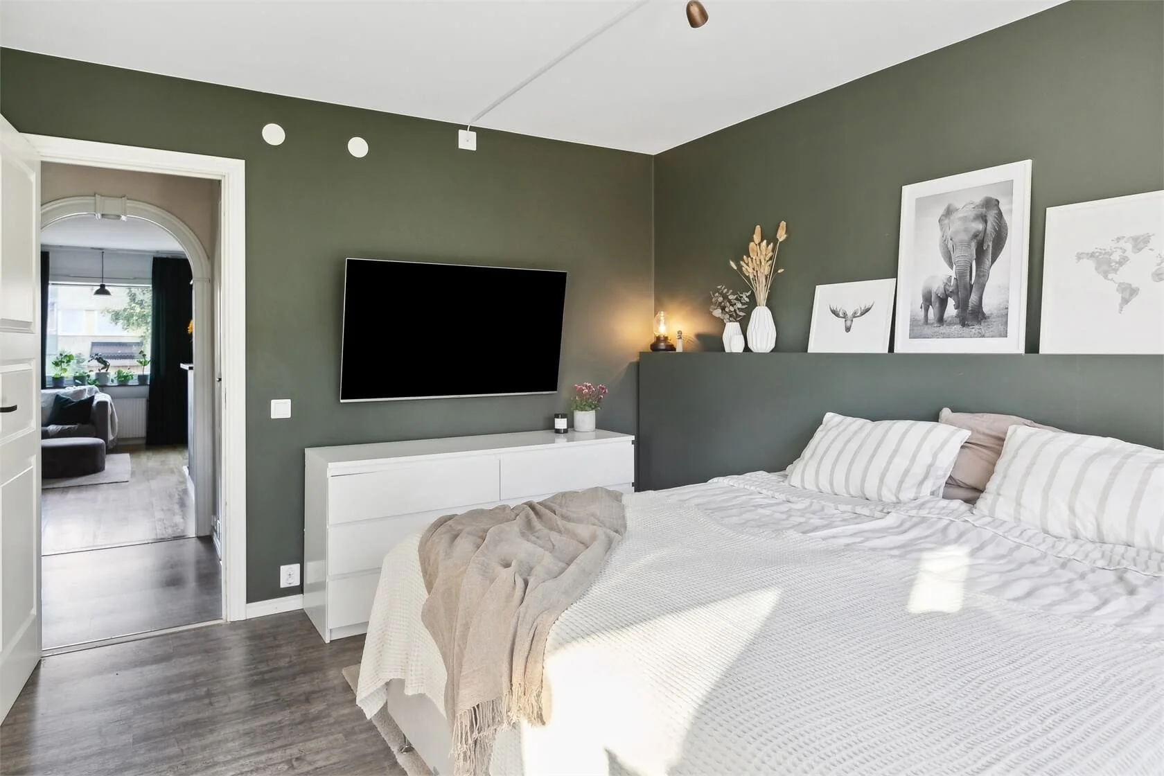 Modern bedroom with dark green wall, white bedding, striped pillows, artwork of animals and world map, wall-mounted TV, and doorway leading to living room with window and plants.