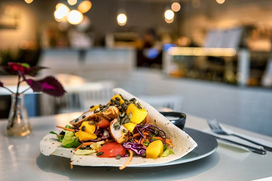Taco filled with sliced chicken, mango, cherry tomatoes, purple cabbage, and greens, topped with pumpkin seeds, on a black plate in a restaurant setting.