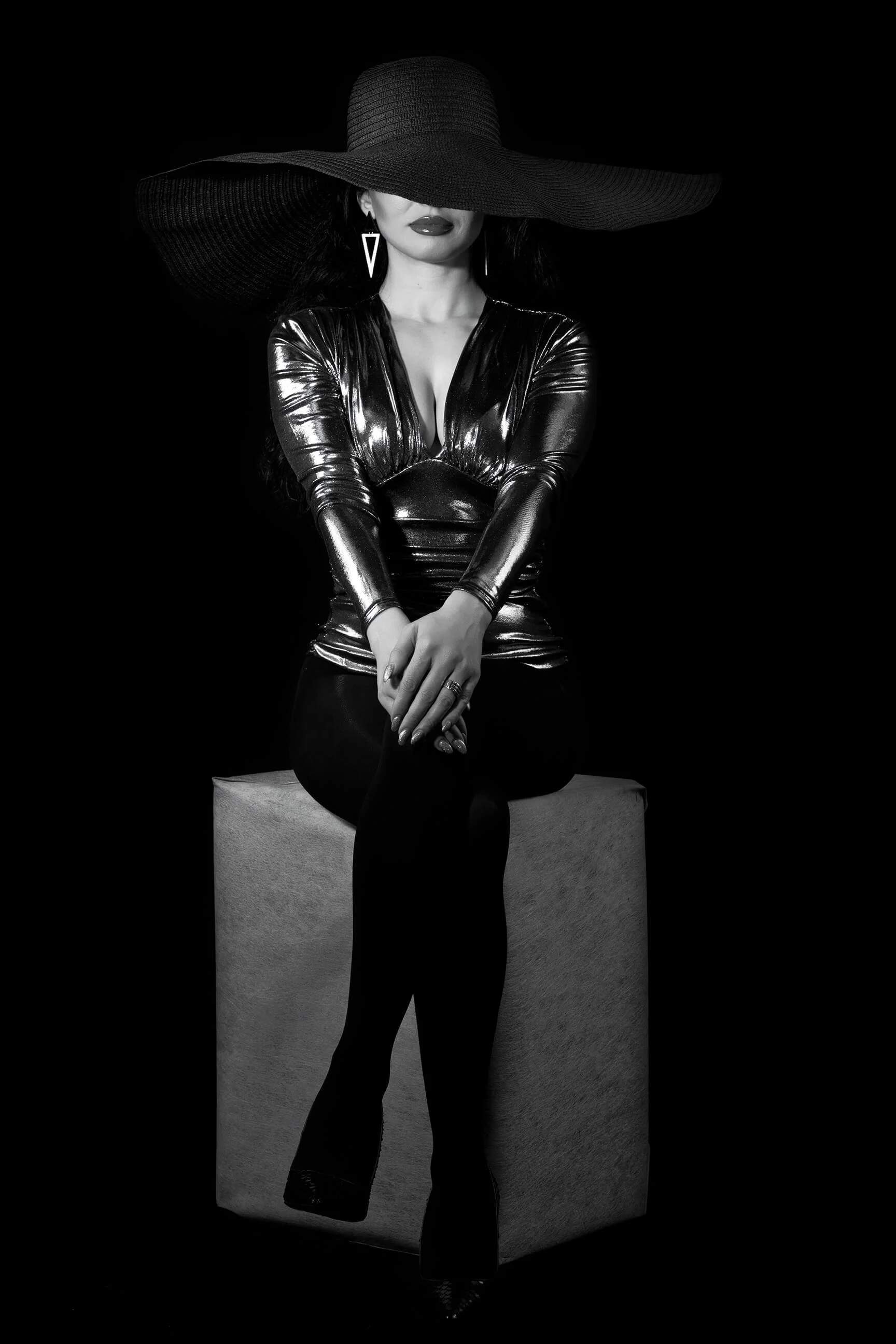 A woman in a shiny, black, form-fitting outfit with long sleeves, sitting on a rectangular block. She wears a very wide-brimmed black hat that obscures part of her face, with lips visible. She has long earrings and rings on her fingers. The image is 