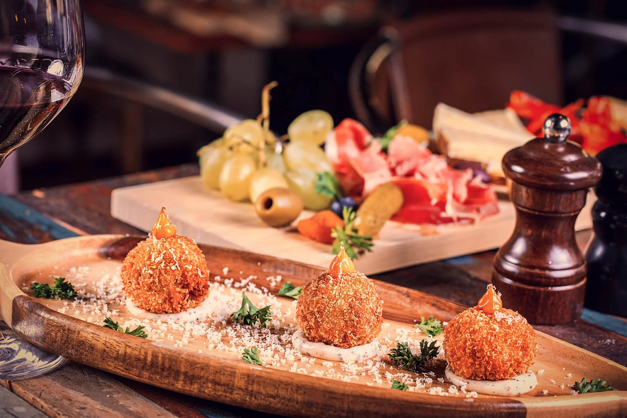 Three breaded and fried appetizer balls topped with a small amount of sauce, served on a wooden platter with chopped herbs and sauce dots, with a charcuterie board of cheese, grapes, and cured meats, and a glass of red wine in the background.